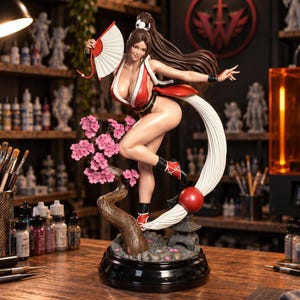 May include: A detailed figurine of a female character with long brown hair, wearing a red and white outfit, holding a white folding fan. The figure stands on a base featuring a brown tree trunk with pink cherry blossoms and a small stone lantern.