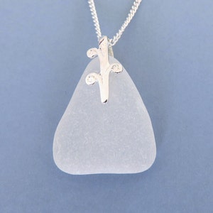 May include: A light blue sea glass pendant necklace with a silver-colored decorative bail and chain. The sea glass is a smooth, triangular shape. The chain is a delicate curb style, and the background is a solid blue.