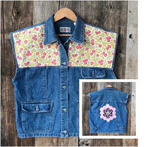 May include: A blue denim vest with a floral print on the collar and shoulders. The vest has a patch pocket on the front and a large floral patch on the back.