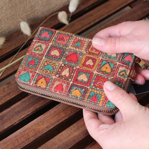 May include: A cork wallet with a zip fastening and a colourful pattern of hearts and squares. The wallet is closed and being held by a hand.