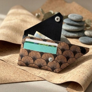 K&ouml;nnte beinhalten: Eine braune und schwarze Korkbrieftasche mit F&auml;chermuster und schwarzem Klappverschluss, der mit silbernen Druckkn&ouml;pfen befestigt ist. Die Brieftasche ist ge&ouml;ffnet und zeigt Kreditkarten und Bargeld. Der Hintergrund zeigt Korkmaterial und einen Stapel grauer Steine.