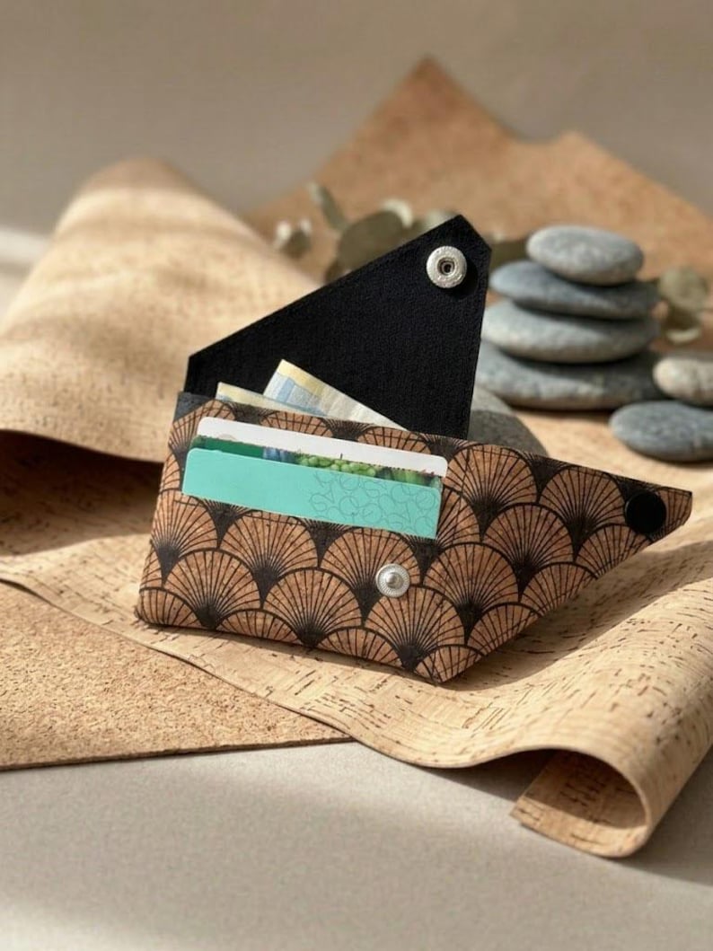 K&ouml;nnte beinhalten: Eine braune und schwarze Korkbrieftasche mit F&auml;chermuster und schwarzem Klappverschluss, der mit silbernen Druckkn&ouml;pfen befestigt ist. Die Brieftasche ist ge&ouml;ffnet und zeigt Kreditkarten und Bargeld. Der Hintergrund zeigt Korkmaterial und einen Stapel grauer Steine.