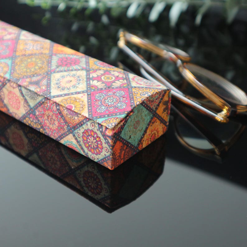 May include: A rectangular patterned glasses case with a colorful geometric design. The case is made of cardboard and has a hinged lid. The case is closed and sitting on a black surface.