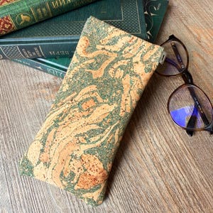 Puede incluir: Un estuche rectangular para gafas con una textura de corcho natural y patrones de remolinos en color tostado y verde bosque. El estuche tiene una forma delgada y alargada con una superficie plana y una pequeña bisagra metálica visible.