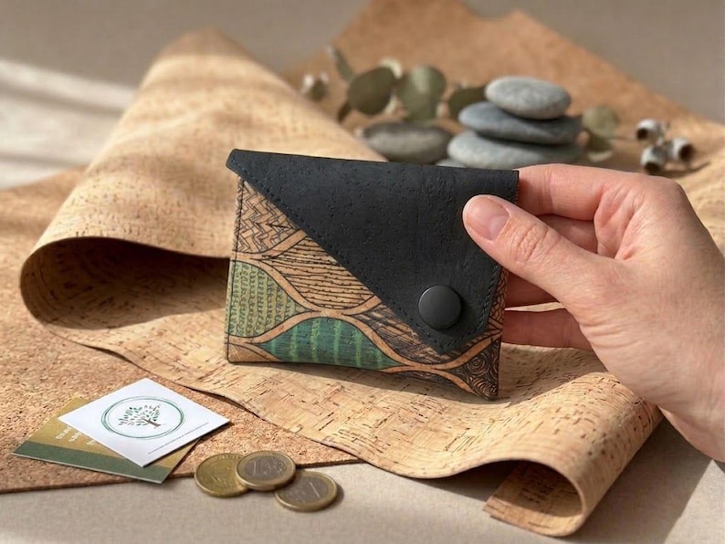 K&ouml;nnte beinhalten: Ein kleines, handgefertigtes Portemonnaie mit schwarzem Klappverschluss und einem gemusterten Korkk&ouml;rper. Das Korkdesign weist gr&uuml;ne und braune wellenartige Muster auf. Das Portemonnaie wird von einer Hand gehalten, und ein kleiner Knopf sichert den Klappverschluss.