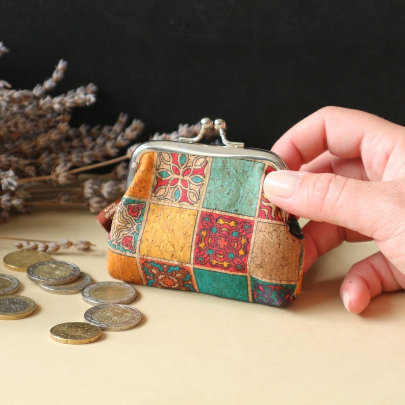 May include: A small, colorful coin purse with a patchwork design. The purse is made of cork and has a silver metal clasp. The purse is sitting on a light brown surface with some coins scattered around it.