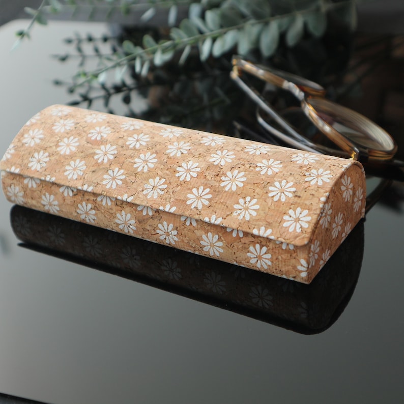May include: A cork and white daisy patterned glasses case. The case is closed and lying on a black surface.