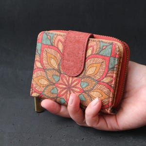 Op de afbeelding: Een kleine rode en bruine kurken portemonnee met een ritssluiting en een bloemmotief mandala. De portemonnee wordt in een hand gehouden.