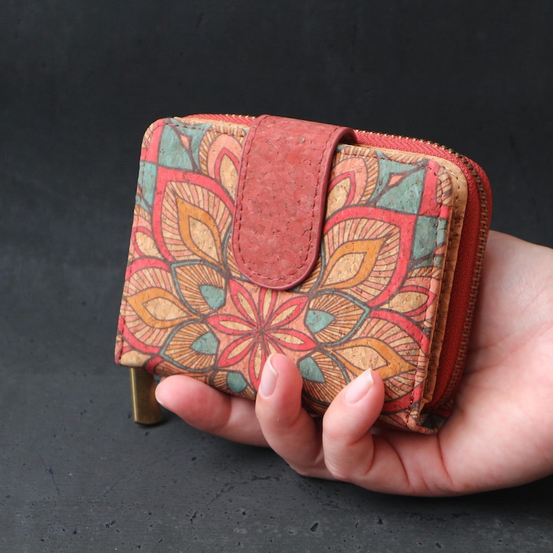 Op de afbeelding: Een kleine rode en bruine kurken portemonnee met een ritssluiting en een bloemmotief mandala. De portemonnee wordt in een hand gehouden.