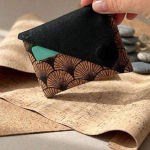 K&ouml;nnte beinhalten: Eine kleine, schwarz-braune Geldb&ouml;rse mit einem f&auml;cherf&ouml;rmigen Design. Die Geldb&ouml;rse ist aus Kork gefertigt und hat einen Druckknopfverschluss. Im Inneren ist eine t&uuml;rkisfarbene Karte sichtbar. Die Geldb&ouml;rse wird in einer Hand gehalten.