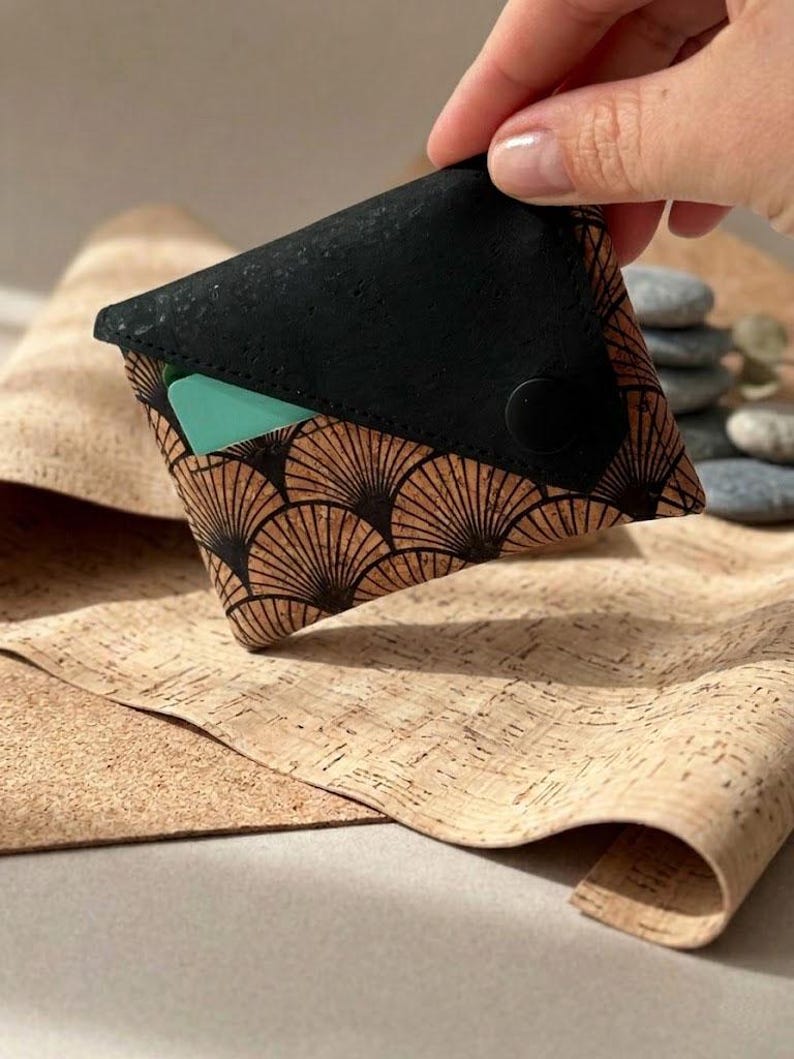 K&ouml;nnte beinhalten: Eine kleine, schwarz-braune Geldb&ouml;rse mit einem f&auml;cherf&ouml;rmigen Design. Die Geldb&ouml;rse ist aus Kork gefertigt und hat einen Druckknopfverschluss. Im Inneren ist eine t&uuml;rkisfarbene Karte sichtbar. Die Geldb&ouml;rse wird in einer Hand gehalten.
