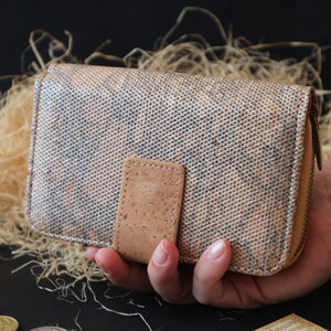 Könnte beinhalten: Eine braune Korkgeldbörse mit Reißverschluss. Die Geldbörse hat ein strukturiertes Muster und einen kleinen Korkfleck auf der Vorderseite.