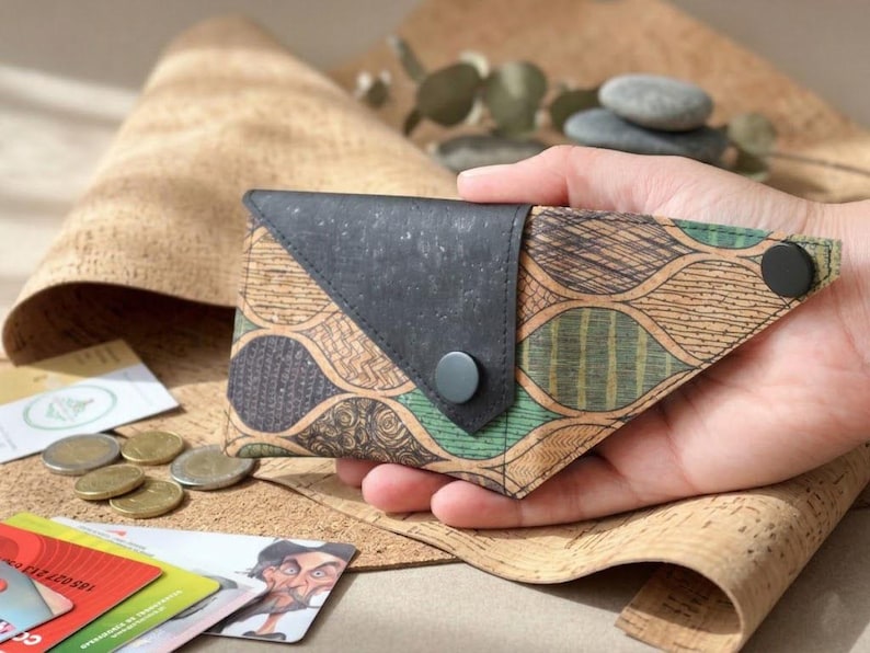 K&ouml;nnte beinhalten: Eine handgefertigte Geldb&ouml;rse mit einem einzigartigen Design. Die Geldb&ouml;rse hat eine schwarze Klappe und einen gemusterten K&ouml;rper mit gr&uuml;nen und braunen Akzenten. Sie wird in einer Hand gehalten, mit M&uuml;nzen und Karten im Hintergrund.