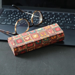 Könnte beinhalten: Ein gemustertes Brillenetui mit einem farbenfrohen Design aus Herzen und Quadraten. Das Etui ist aus Pappe und hat einen braunen Hintergrund mit roten, orangen, gelben, grünen und blauen Akzenten.