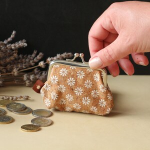 May include: A small cork purse with a white floral pattern and a silver clasp. The purse is open and a hand is reaching into it. There are coins scattered on the table in front of the purse.
