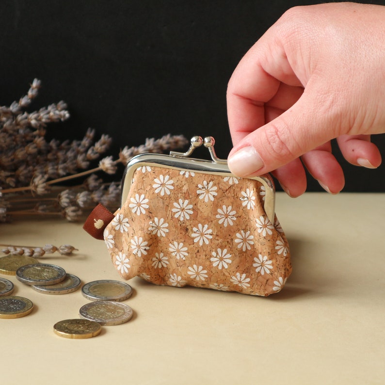 May include: A small cork purse with a white floral pattern and a silver clasp. The purse is open and a hand is reaching into it. There are coins scattered on the table in front of the purse.