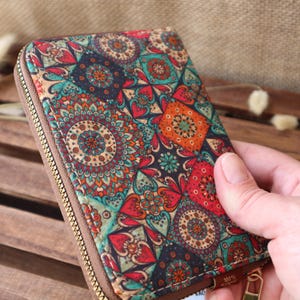 May include: A small zippered wallet made of cork with a colourful mandala pattern. The wallet has a zipper closure and a brown leather trim. The text "100%" is visible on the bottom right corner of the wallet.