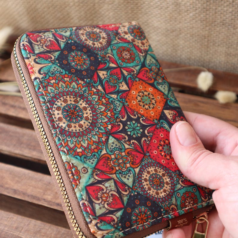May include: A small zippered wallet made of cork with a colourful mandala pattern. The wallet has a zipper closure and a brown leather trim. The text "100%" is visible on the bottom right corner of the wallet.