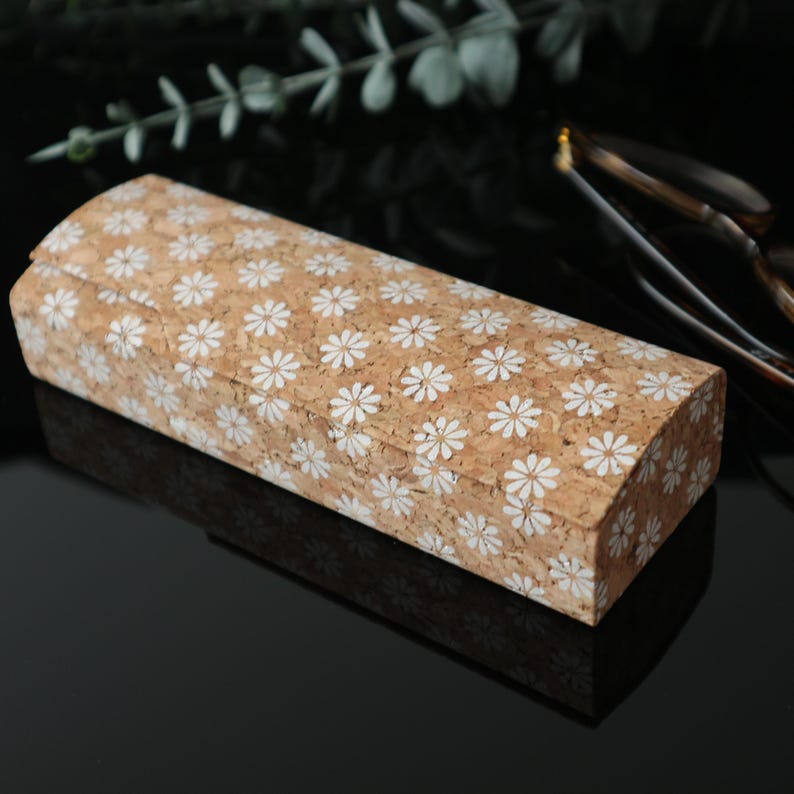 May include: A rectangular cork eyeglasses case with a white daisy pattern. The case is closed and sitting on a black surface.