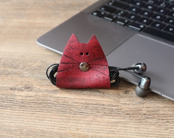 May include: A red cork leather cat-shaped headphone holder with black stitching and a silver button. The holder is holding a pair of black headphones with silver ear buds.