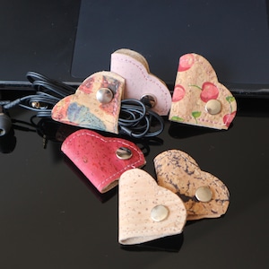 May include: A selection of heart-shaped cork earbud tidies in different colours and patterns. One is pink, another red, and others feature cherry and abstract designs. Each has a silver snap closure.