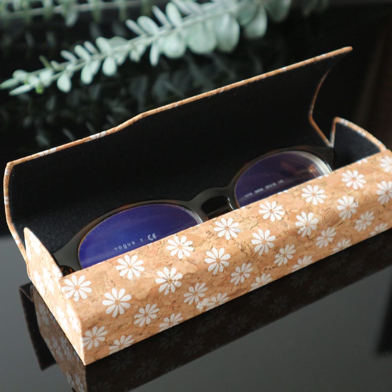 May include: A cork and fabric eyeglasses case with a white daisy pattern. The case is open and a pair of eyeglasses with blue lenses is inside. The text "Vogue T CE" is visible on the glasses.