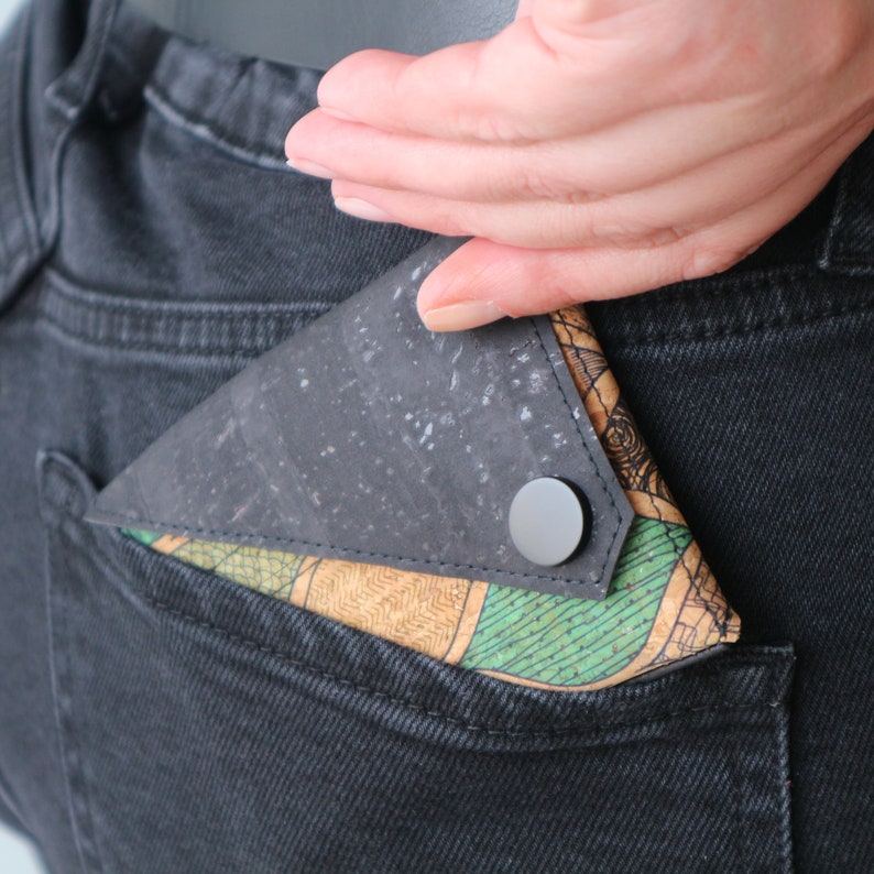 K&ouml;nnte beinhalten: Ein schwarzes Portemonnaie mit einem gesprenkelten Muster und einem Druckknopfverschluss ist teilweise in einer Tasche zu sehen. Die Innenseite hat einen gr&uuml;n-beigen Stoff mit Muster. Das Portemonnaie wird in eine schwarze Jeans-Tasche gesteckt.