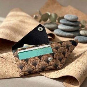 K&ouml;nnte beinhalten: Eine braune und schwarze Korkbrieftasche mit einem f&auml;cherartigen Muster, ge&ouml;ffnet, um Kreditkarten zu zeigen. Die Brieftasche wird mit einem Druckknopf verschlossen. Der Hintergrund zeigt Korkmaterial und einen Stapel grauer Steine.