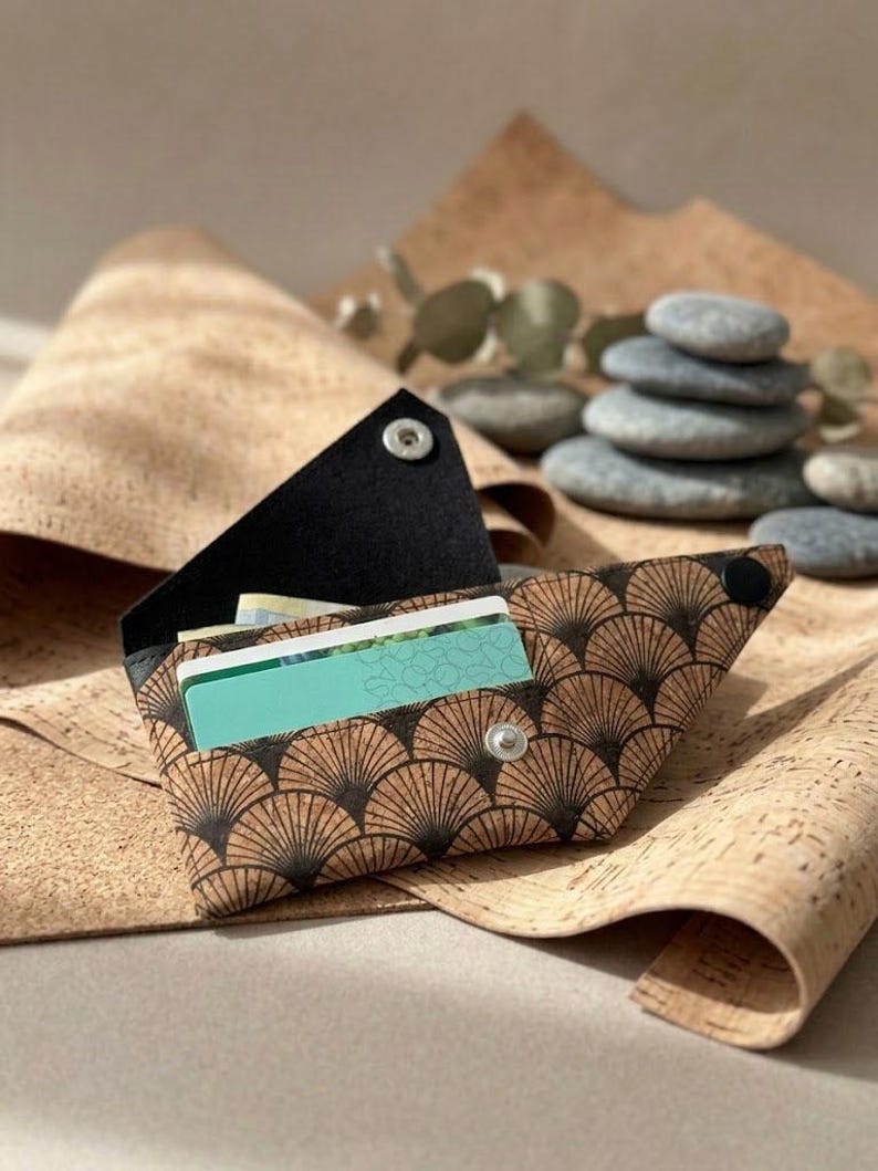 K&ouml;nnte beinhalten: Eine braune und schwarze Korkbrieftasche mit einem f&auml;cherartigen Muster, ge&ouml;ffnet, um Kreditkarten zu zeigen. Die Brieftasche wird mit einem Druckknopf verschlossen. Der Hintergrund zeigt Korkmaterial und einen Stapel grauer Steine.