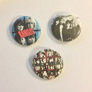 May include: Three round pinback buttons featuring the band The Bangles. The first button has a blue background with a red band across the middle that says "Bangles". The second button has a black and white photo of the band members. The third button has a grid of black and white photos of the band members.