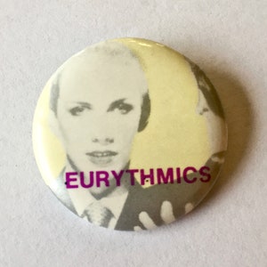 May include: A round button with a black and white photo of a woman with short hair. The text "EURYTHMICS" is printed in purple on the button.