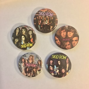 May include: Five round Skid Row band buttons. Each button features a different image of the band members, with the band's name in a stylized font. The buttons have a glossy finish and are arranged in a circular pattern.