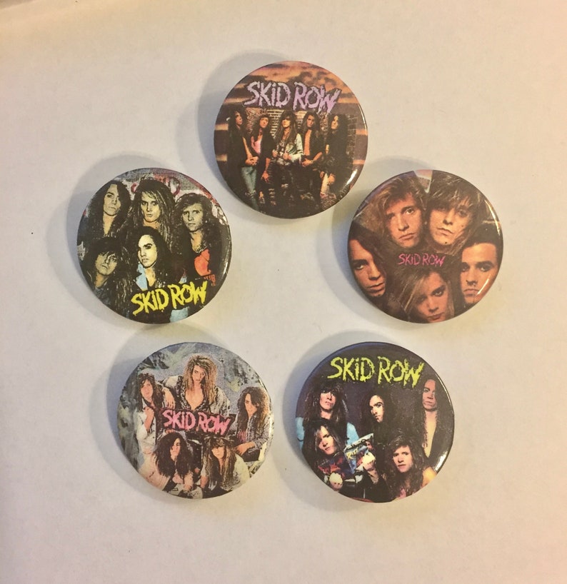 May include: Five round Skid Row band buttons. Each button features a different image of the band members, with the band's name in a stylized font. The buttons have a glossy finish and are arranged in a circular pattern.