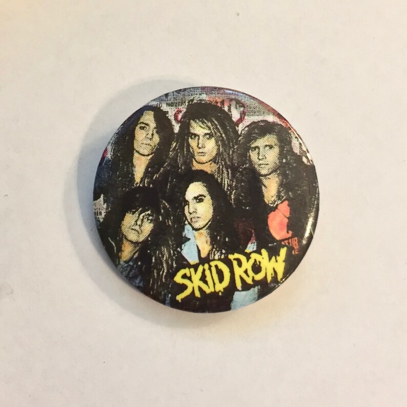 May include: A round button featuring a band portrait. The button has a dark background with the band members' faces and the band name "SKID ROW" in yellow letters.