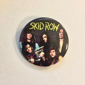 May include: A round button featuring the band Skid Row. The button has a dark background with the band members' faces and the band's name in yellow, stylized text. The button is a collectible item.
