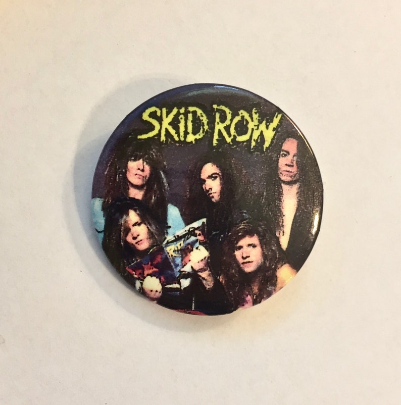 May include: A round button featuring the band Skid Row. The button has a dark background with the band members' faces and the band's name in yellow, stylized text. The button is a collectible item.