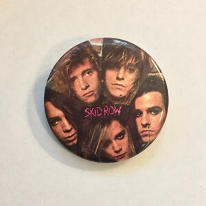 May include: A round button featuring a collage of five faces, with the band name "SKID ROW" in pink text. The button has a glossy finish and is set against a white background.