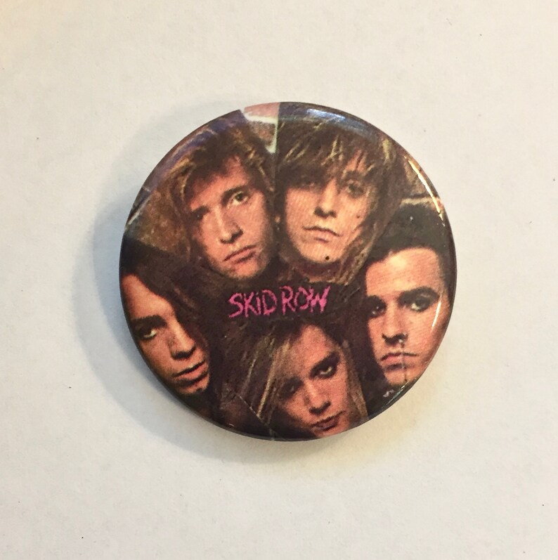 May include: A round button featuring a collage of five faces, with the band name "SKID ROW" in pink text. The button has a glossy finish and is set against a white background.