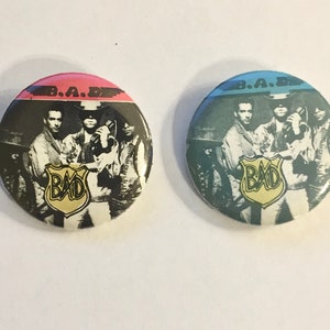May include: Two round pinback buttons with a black and white image of a band called "BAD" with the text "B.A.D." above the image. One button has a pink background and the other has a blue background.