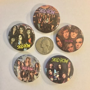 May include: A collection of five Skid Row band buttons. Each button features a different image of the band members, with the band's name prominently displayed. The buttons are round and have a glossy finish. A quarter is included for size comparison.