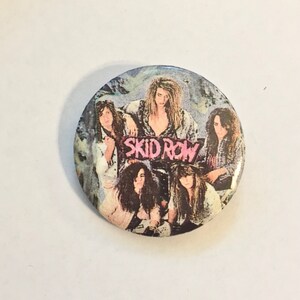 May include: A round button featuring the band Skid Row. The button has a group photo of the band members with long hair and a grunge aesthetic. The band's name is in pink letters.