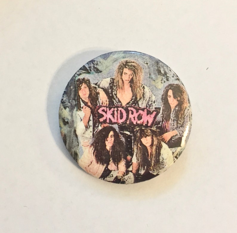 May include: A round button featuring the band Skid Row. The button has a group photo of the band members with long hair and a grunge aesthetic. The band's name is in pink letters.
