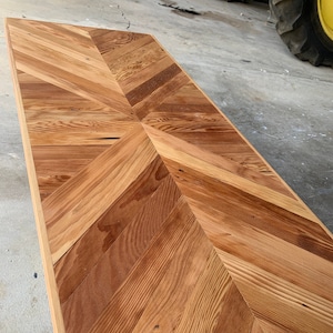 Puede incluir: Una tapa de mesa de madera con un patrón de espiga hecho de tablones de madera recuperados. La madera tiene un color marrón natural con variaciones en la veta y la textura.