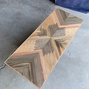 May include: A rectangular coffee table with a geometric pattern made of different types of wood. The table has a light brown border and a dark brown centre with a light brown starburst design.