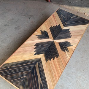 May include: Wooden rectangular table with a geometric design. The table has a central leaf-like pattern in black, surrounded by natural wood tones. The table is made of wood and has a smooth finish.