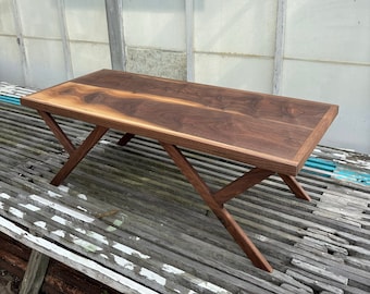 Puede incluir: Una mesa de centro de madera marrón oscuro con una tapa rectangular y una base en forma de X. La mesa está hecha de madera de nogal y tiene un acabado natural.