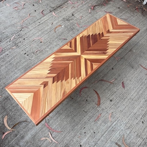 Puede incluir: Una mesa rectangular de madera con un diseño geométrico. La tapa presenta un patrón central en forma de cruz y un borde, todo elaborado con varias tonalidades de madera. La mesa mide aproximadamente 152 cm de largo.