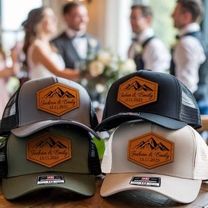 May include: Four trucker hats in gray, black, olive green, and tan, each with a mesh back and a leatherette patch. The patch features a mountain design and the text "Jackson & Emily 15.11.2025".