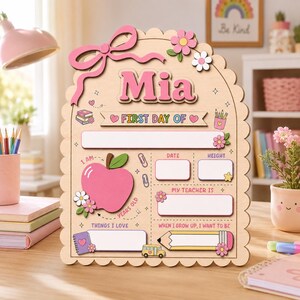 Puede incluir: Un letrero de madera festoneado para el primer d&iacute;a de clases con el nombre Mia en letras rosas. El tablero incluye secciones para fecha, altura, profesor, edad, gustos y profesi&oacute;n, decorado con una manzana rosa, flores y un l&aacute;piz.
