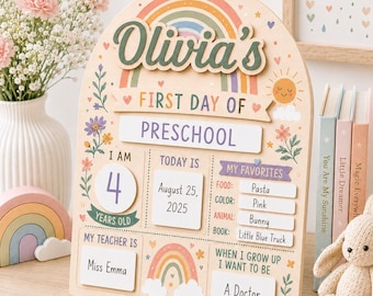 May include: A light-colored wooden arch-shaped sign featuring a rainbow graphic and the text Olivia's First Day of Preschool. The board includes sections for age, date, teacher, favorites, and future goals, decorated with flowers and a sun.